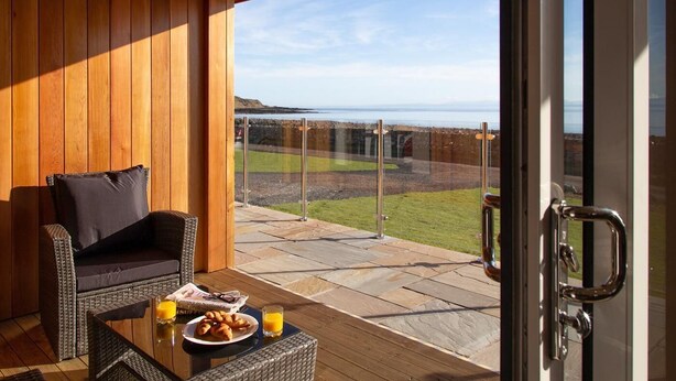 Een balkon met een rieten bank, een glazen tafel met ontbijtartikelen en uitzicht op zee.