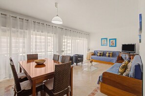 A modern living room with a dining area, a sofa set, a television, and artwork on the wall. - Apartment 'Estrela Do Forte' with Shared Pool, Balcony and Wi-Fi (Quarteira)