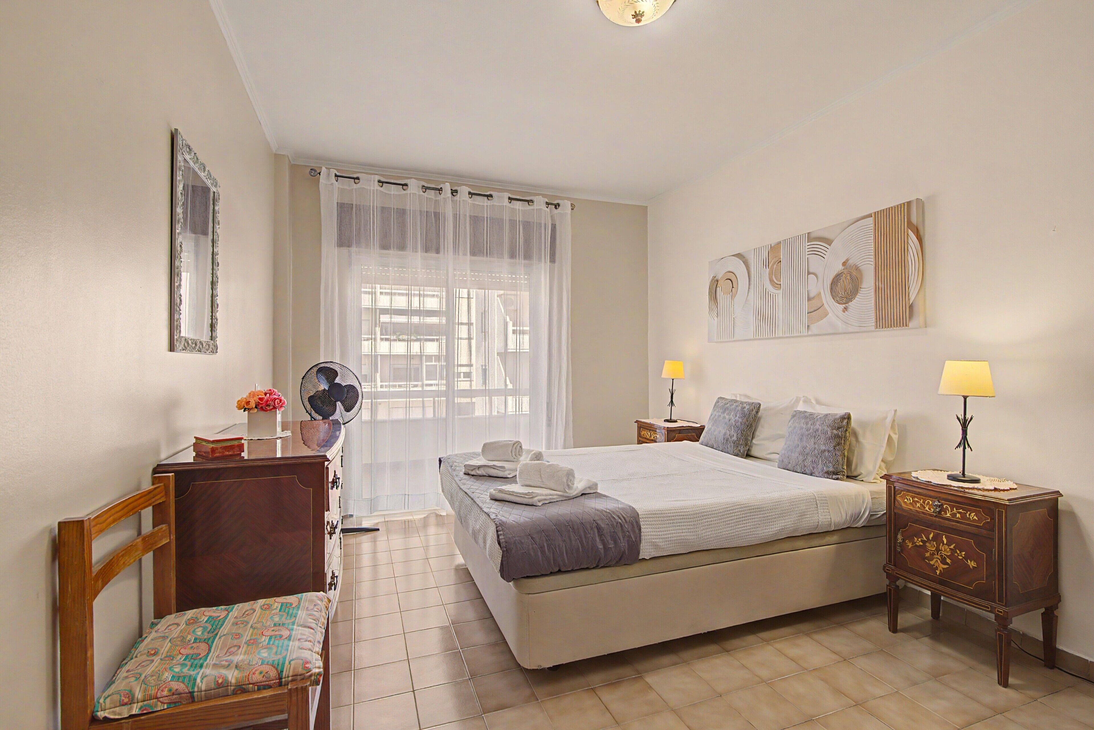 A hotel room with a bed, a desk, a chair, a bedside table with a lamp, a wall-mounted artwork, and a window with sheer curtains.