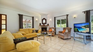 Un salón con sofá amarillo, chimenea, televisor y vistas al exterior.