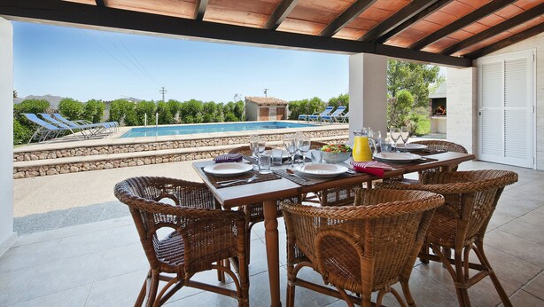 Un espacio comedor al aire libre con techo, sillas de mimbre y una mesa dispuesta para dos personas, con vistas a una piscina y montañas al fondo.