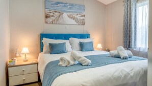 A neatly made bed with blue bedding, a bedside table with a lamp, and a painting of a beach scene on the wall. - 2 Bed in Upton (oc-c35149) (Ryde)