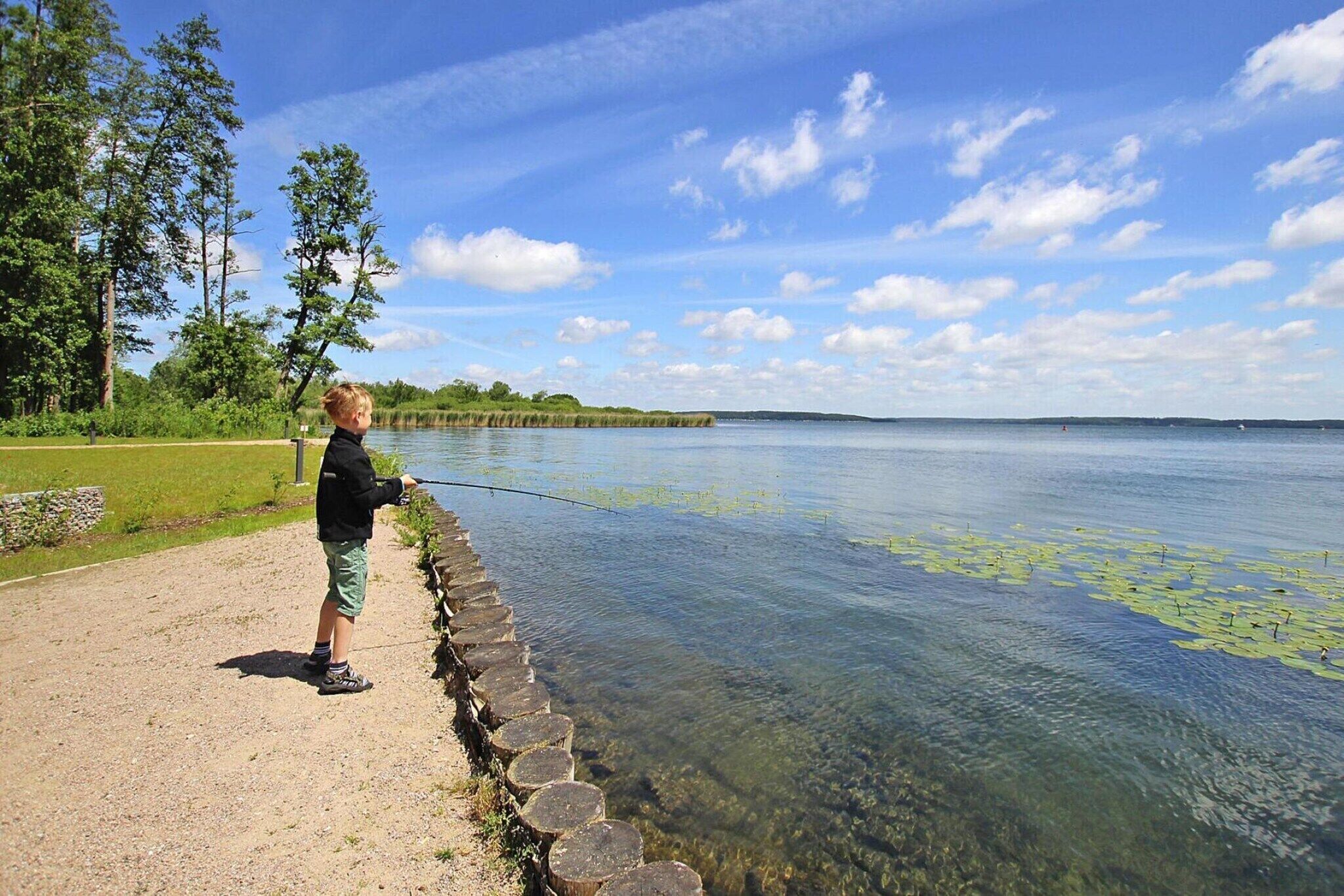 A child fishing by a calm lake with clear water, surrounded by trees and a stone path.