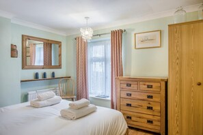 A bedroom with a bed, a wooden dresser, a chair, a radiator, a window with curtains, a chandelier, and a framed picture on the wall.