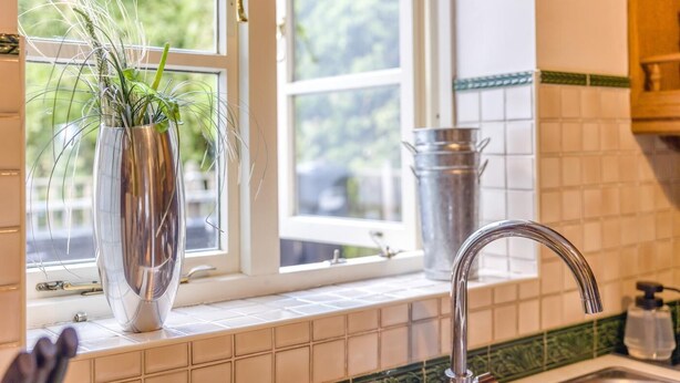 Una cocina con ventana, un florero con plantas, un recipiente metálico y un grifo.