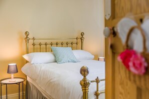 Una cama con cabecera de metal, ropa de cama blanca y una sola almohada decorativa. Hay una lámpara de noche sobre una mesita junto a la cama.