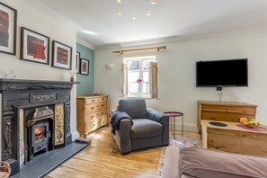 Ein Wohnzimmer mit Kamin, einem grauen Sessel, einem Fernseher und einer Holzkommode.