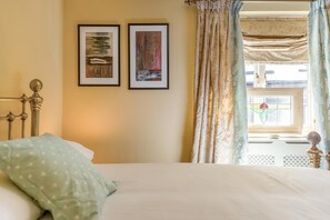 Ein Schlafzimmer mit Bett, Kopfteil, zwei gerahmten Kunstwerken, einem Fenster mit Vorhängen und Blick ins Freie.