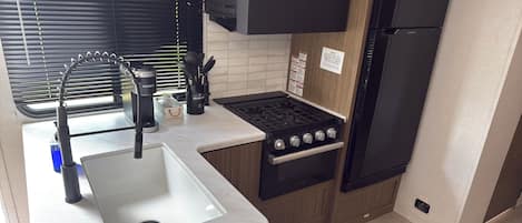 Une cuisine compacte équipée d’un évier, d’une cuisinière et d’un réfrigérateur, avec une fenêtre dotée de stores et un petit tapis.