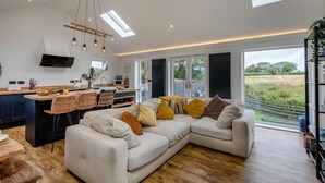 Una sala de estar moderna con un sofá modular, un comedor con mesa y sillas, una cocina con televisor de pantalla plana y ventanas amplias con vistas al exterior.