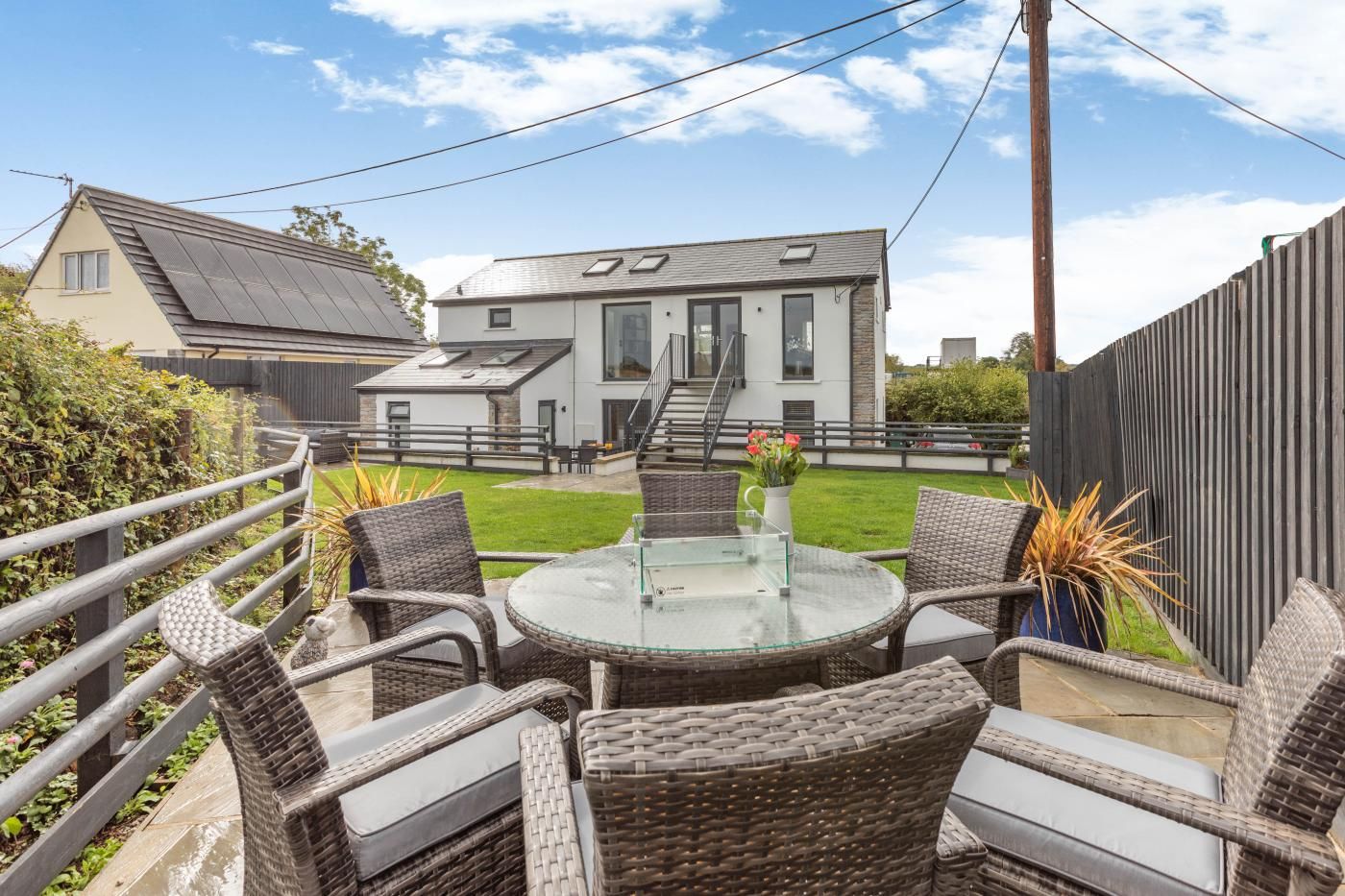 A patio with wicker furniture and a glass table, a modern house in the background, and a wooden fence.