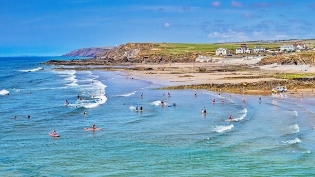 Escena de playa costera con olas, gente y casas al fondo.