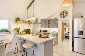 A modern kitchen with a central island, bar stools, a built-in oven, and a large clock on the wall. - 2 Bed in Lanteglos (oc-l35091) (Lanteglos Highway)