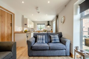 A modern living room with a grey sofa, a wooden door, and a kitchen in the background. - 1 Bed in Child Okeford (oc-f34999) (Child Okeford)