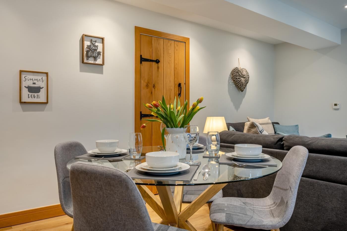 A dining area with a glass table, grey upholstered chairs, a wooden door, and a wall-mounted light fixture.
