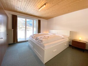 A bedroom with a large bed, a wooden headboard, a nightstand with a lamp, and a view of mountains through the window. - Haus Montana 3/7 Appartement/Fewo,   5-Bettwohnung by Interhome (Lenzerheide)