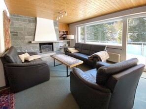 Ein modernes Wohnzimmer mit Kamin aus Stein, Ledersofas, einem Holztisch und großen Fenstern mit Blick auf Bäume.