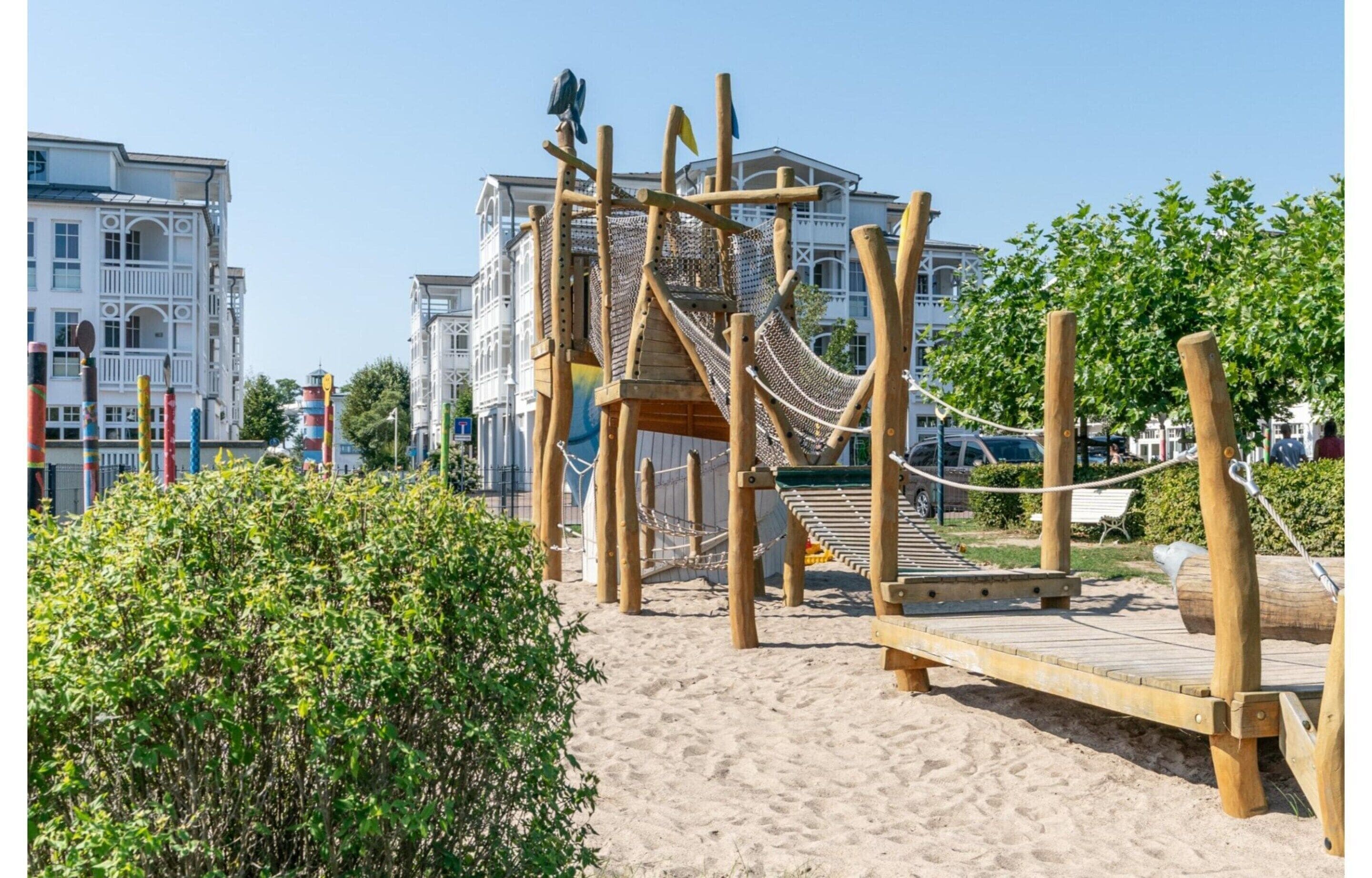 A wooden playground with climbing structures and a slide, surrounded by sand and greenery, with buildings in the background.