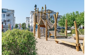 A wooden playground with climbing structures and a slide, surrounded by sand and greenery, with buildings in the background. - Komfortable Ferienwohnung im Seepark Sellin (Sellin)