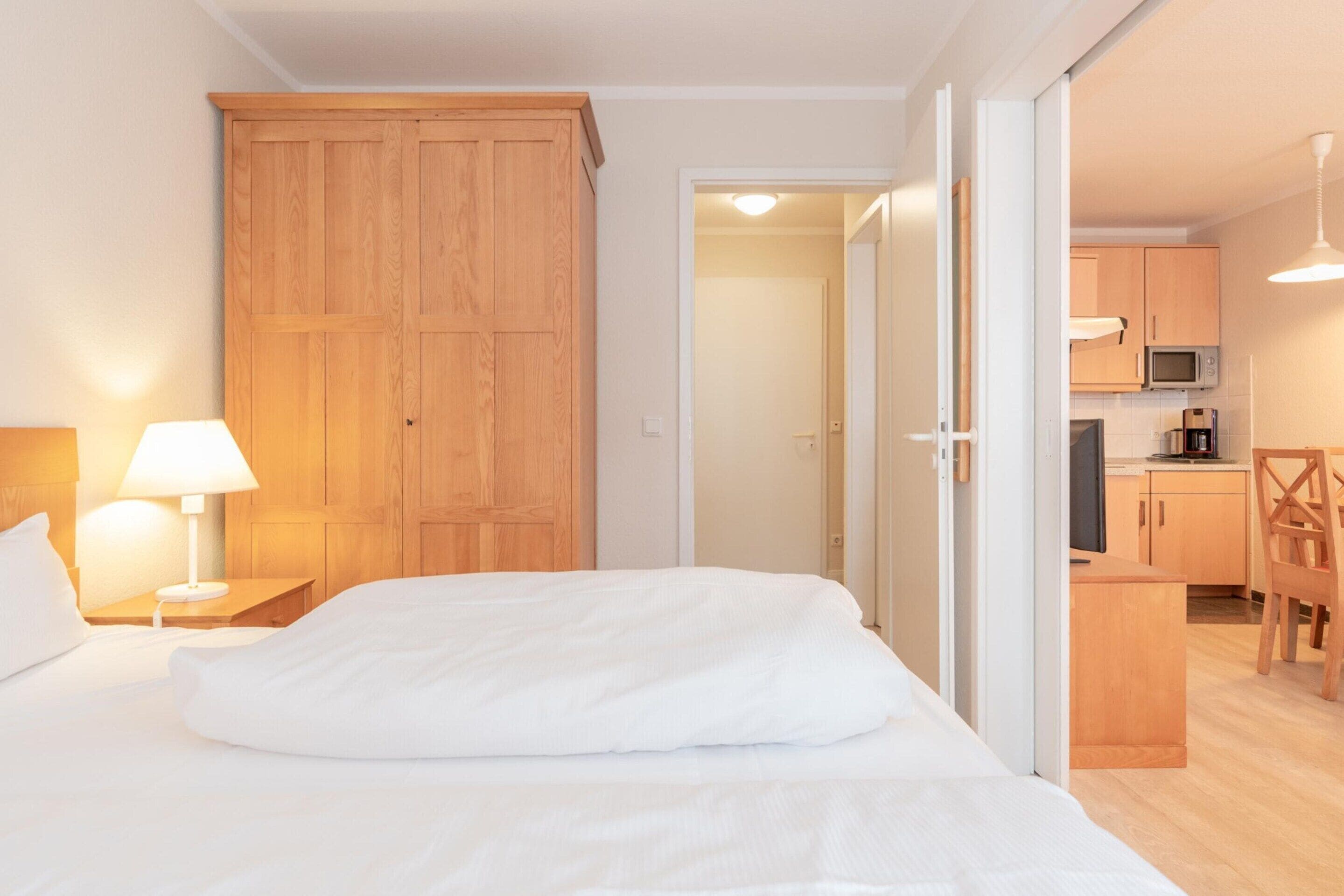 Une chambre d’hôtel avec un lit, une armoire en bois, un bureau avec une lampe et, au fond, une petite cuisine.