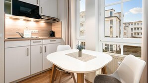 Eine moderne Küche mit weißen Schränken, einer Mikrowelle und einer Kaffeemaschine. Ein Esstisch mit Stühlen bietet Blick auf Gebäude durch große Fenster.