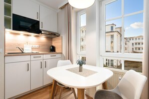 Eine moderne Küche mit weißen Schränken, einer Mikrowelle und einer Kaffeemaschine. Ein Esstisch mit Stühlen bietet Blick auf Gebäude durch große Fenster.