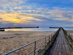 A wooden boardwalk leading to a pier with a lighthouse, a sandy beach, and a calm sea under a partly cloudy sky. - 5 Bed in Blyth (oc-t34727) (Blyth)