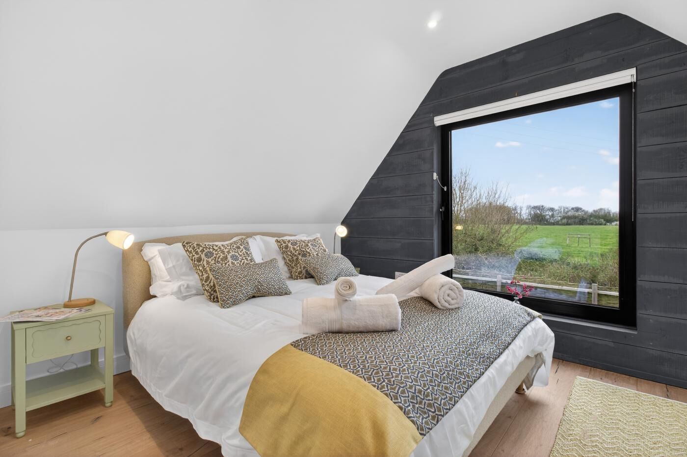 Une chambre avec une grande fenêtre, un lit recouvert d’une literie à motifs, une table de chevet avec une lampe, et une vue sur un paysage verdoyant.