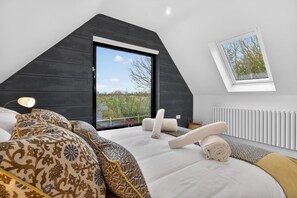 A bedroom with a sloped ceiling, a large window, a bed with patterned bedding, and a bedside table.