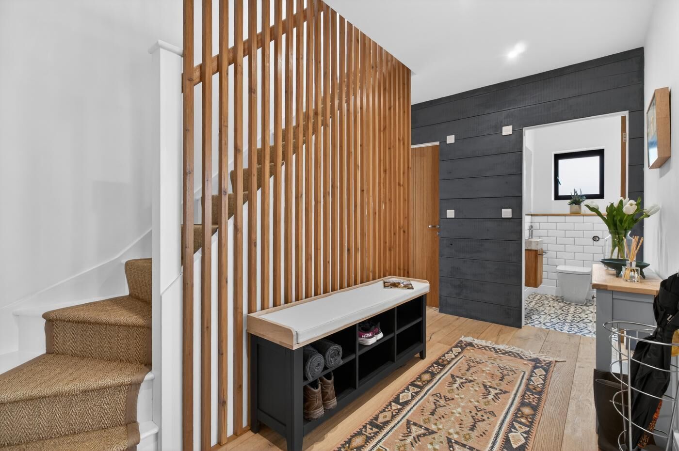 Un intérieur moderne avec un escalier, une cloison en bois, un banc avec espace de rangement, un tapis à motifs et une petite plante.
