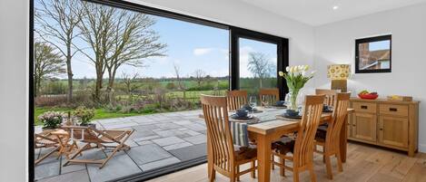 A dining area with a table set for four, wooden chairs, a wooden cabinet, and a view of the outdoors.