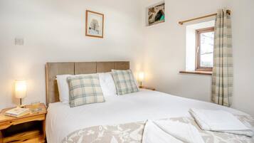 A neatly made bed with plaid pillows, a bedside table with a lamp, a framed picture on the wall, and a window with curtains.