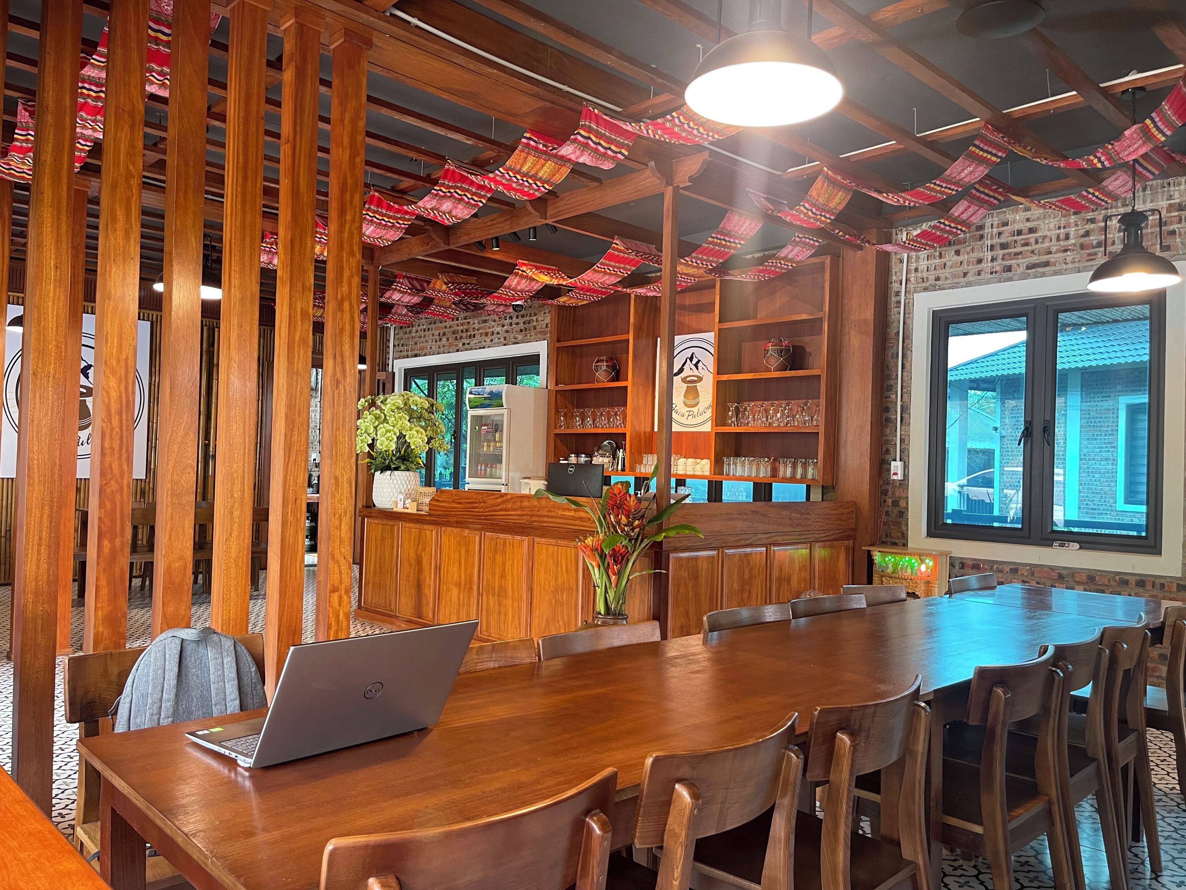 A wooden interior with a long table and chairs, a laptop on the table, and a brick wall with a window.