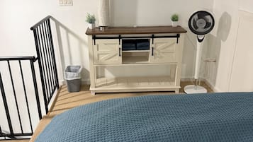 Et soveværelse med seng, kommode, en lille plante, ventilator og et vindue.