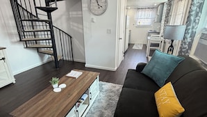 A living room with a black sofa, a wooden coffee table, a spiral staircase, and a clock on the wall. - Cozy Creek Retreat - Spiral staircase,AC,high speed WiFi (Waterford)