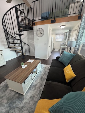 Ein Wohnzimmer mit einer schwarzen Couch, einem Holztisch, einer Wendeltreppe und einer Uhr an der Wand.