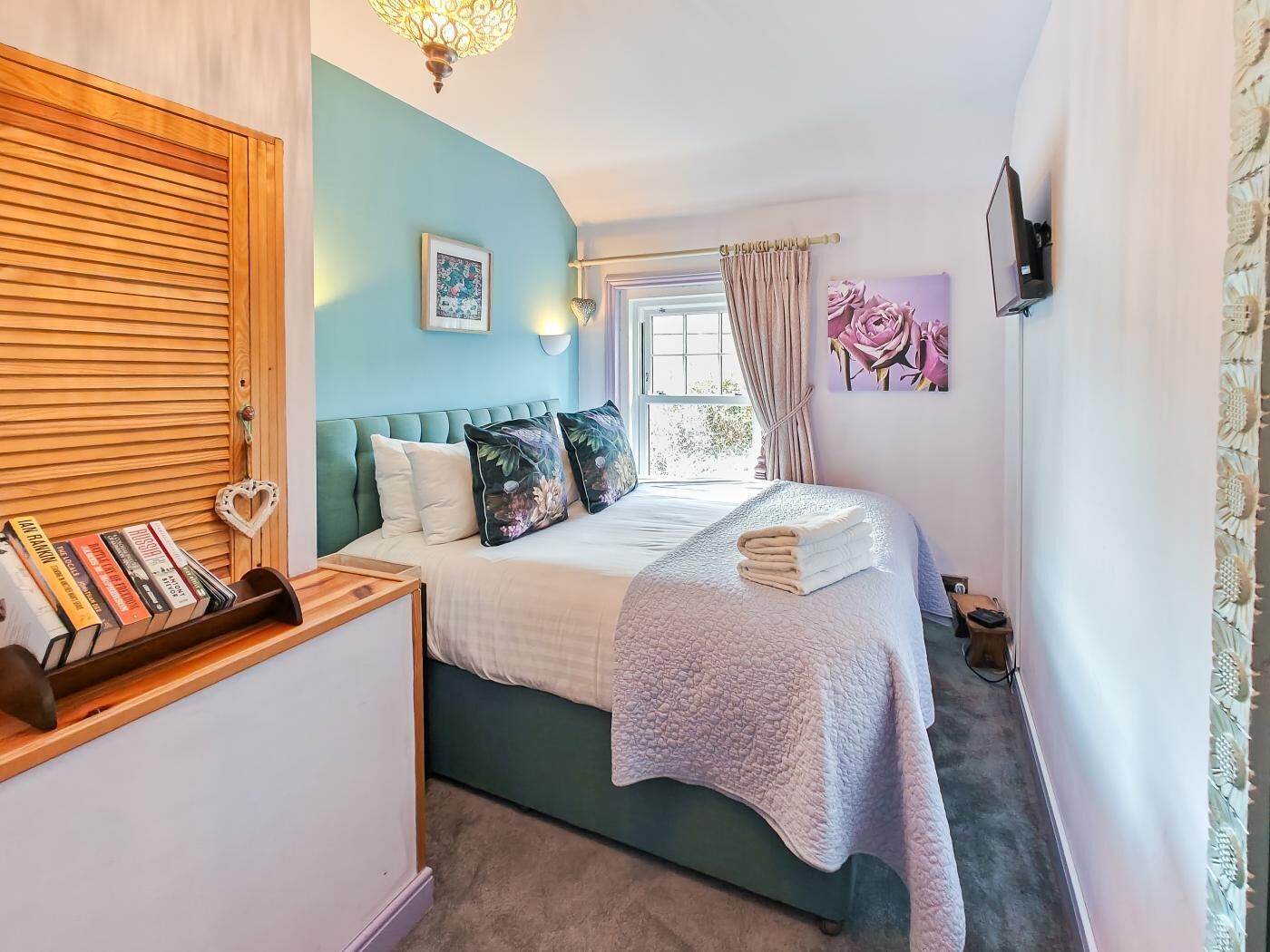 A bedroom with a bed, a bedside table with books, a window with curtains, and a wall-mounted TV.