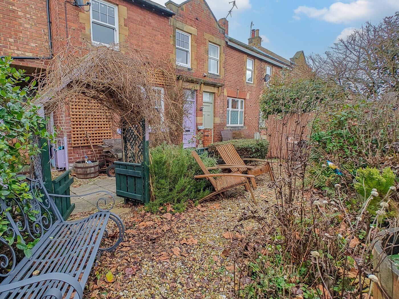 A residential garden with a wooden bench, a green metal gate, and a brick house with multiple windows.