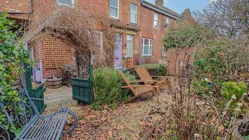A residential garden with a wooden bench, a green metal gate, and a brick house with multiple windows.