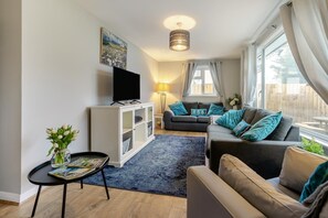 Ein Wohnzimmer mit einer Couch, einem Fernseher auf einem Schrank, einer Lampe und einem Fenster mit Vorhängen.