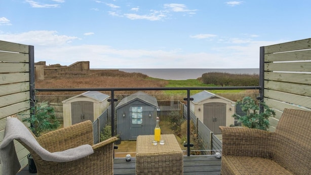 Ein Balkon mit Korbmöbeln, einem Tisch mit einem Glas Saft und Blick auf das Meer und Strandhäuser.