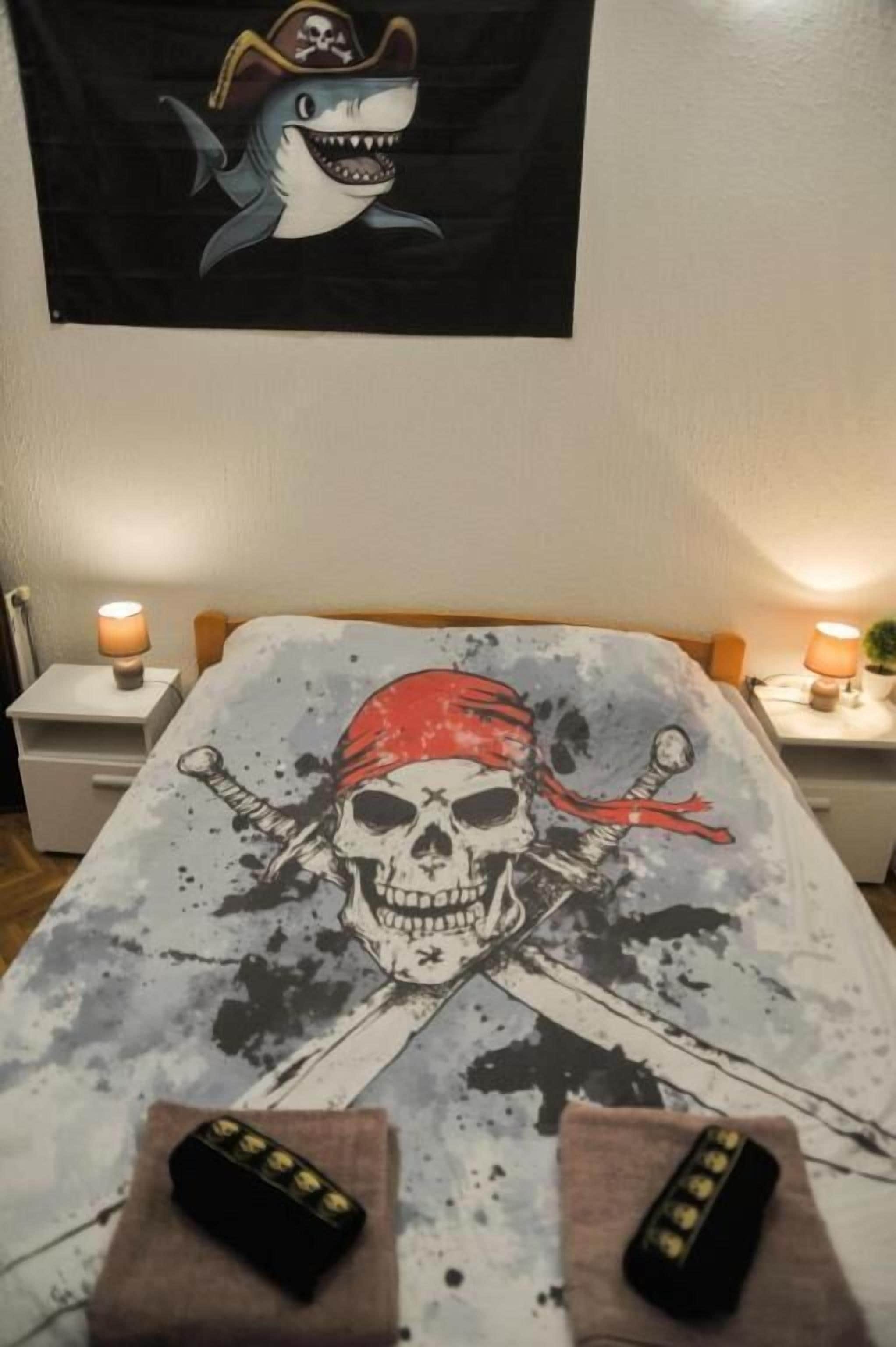 Un lit avec une couvre-lit sur le thème des pirates, deux télécommandes posées sur le lit et un mur décoré d’un drapeau représentant un requin et un autre représentant des pirates.