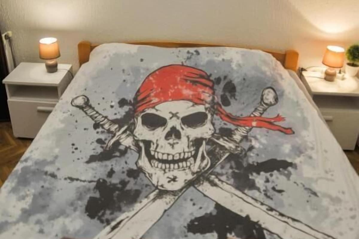 A bed with a pirate-themed bedspread, two remote controls on the bed, and a wall with a shark and pirate flag.
