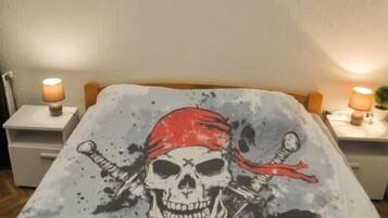 Un lit avec une couvre-lit sur le thème des pirates, deux télécommandes posées sur le lit et un mur décoré d’un drapeau représentant un requin et un autre représentant des pirates.