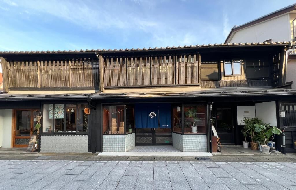 Un bâtiment japonais traditionnel avec une façade en bois, un auvent bleu et un petit espace extérieur aménagé avec des plantes.