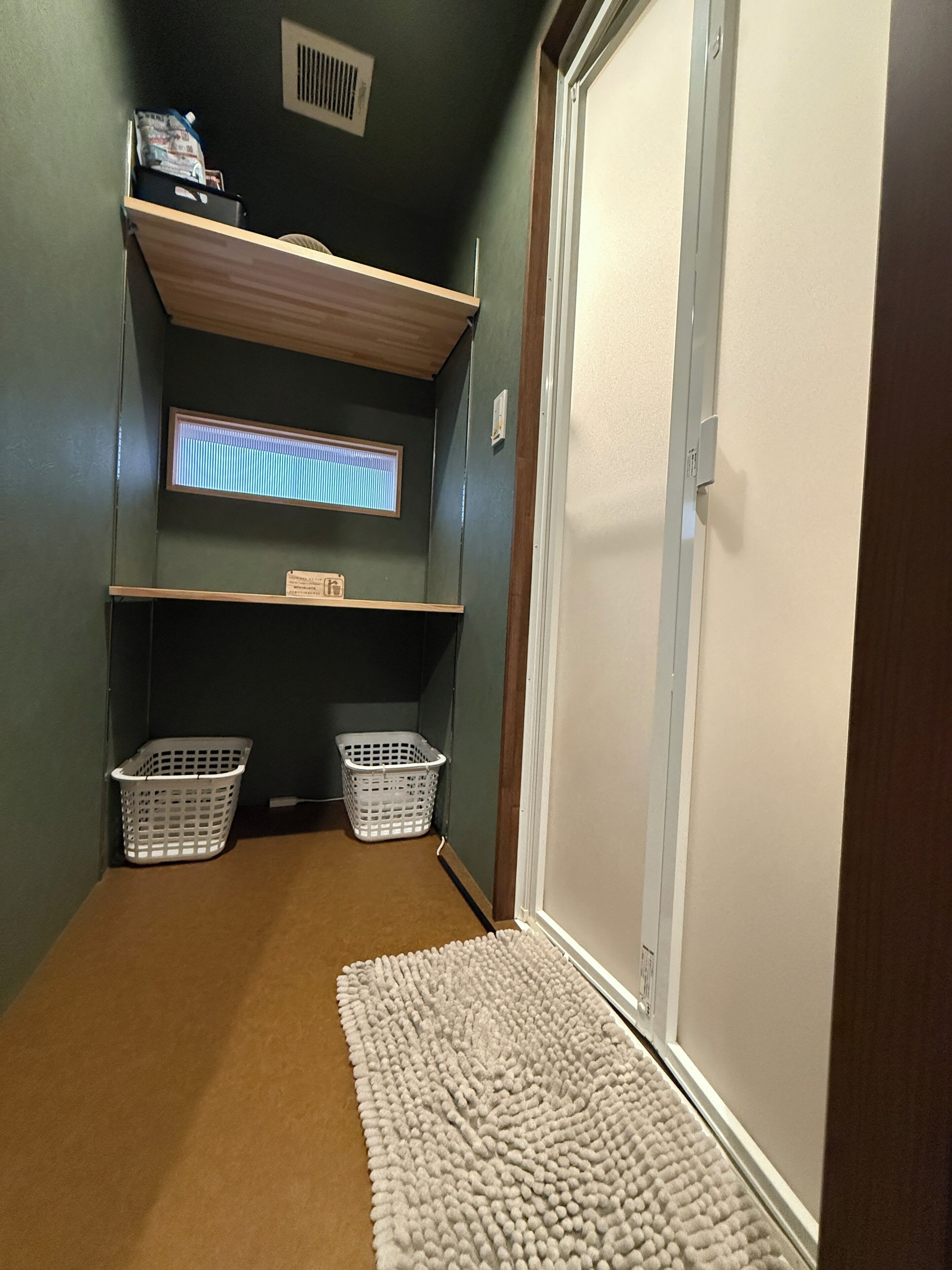 A hallway with a carpeted floor, two white baskets, a shelf with items, a vent, and a door.