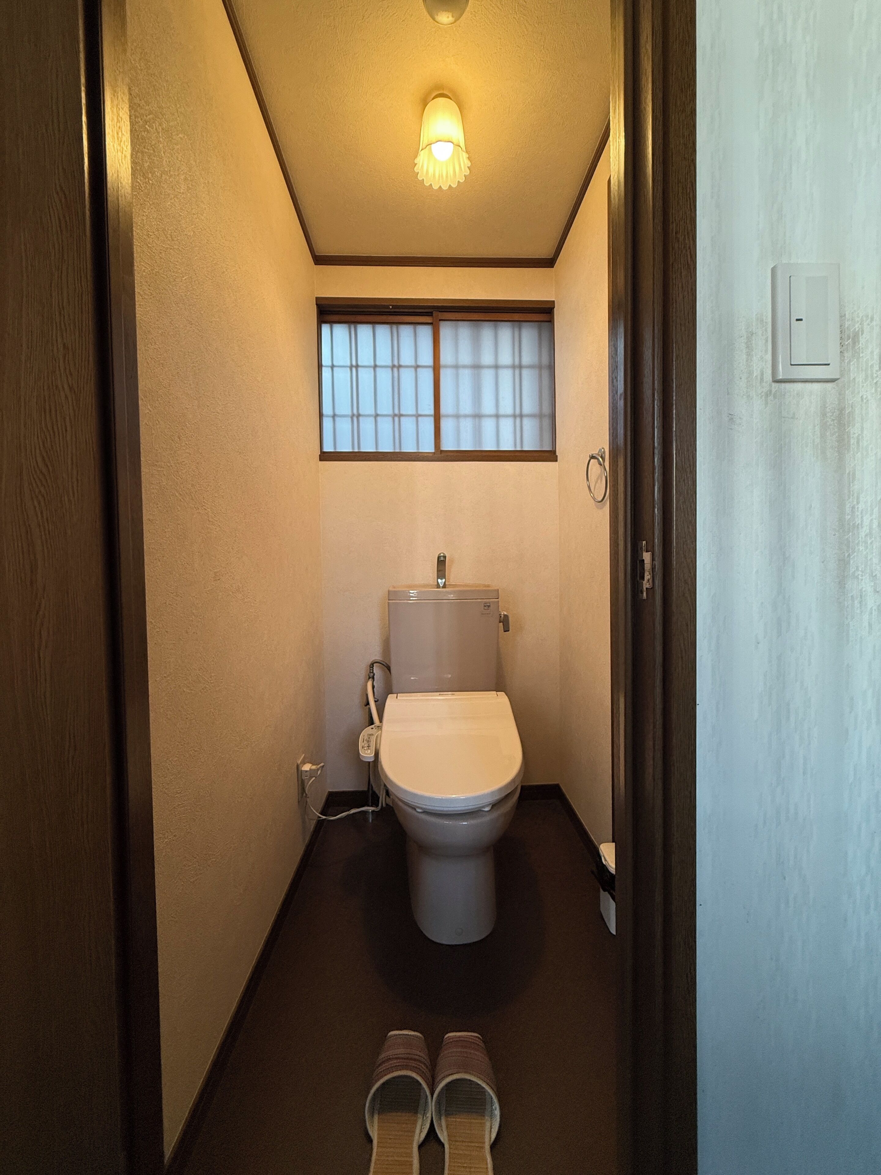 A small, narrow bathroom with a toilet, a window, and a pair of slippers on the floor.