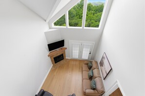 A room with a wooden floor, a sofa, a TV, a fireplace, and a large window with a view of trees. - Spacious 3-bedroom condo with River and Mountain Views (Sevierville)