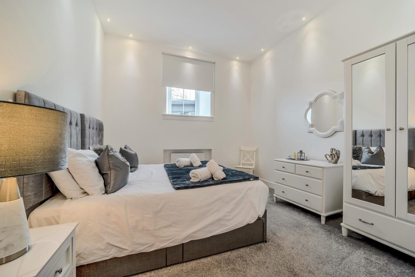 Une chambre à coucher comprenant un lit, une table de chevet, une lampe, une commode, un miroir et une fenêtre.