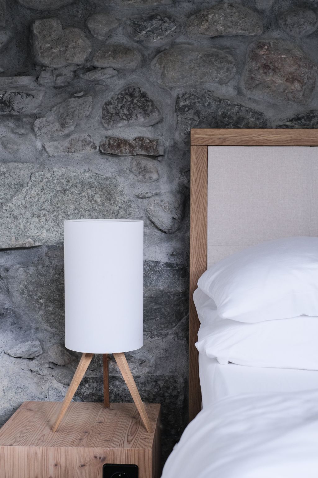 A bed with white linens against a stone wall, a bedside table with a lamp, and a wooden nightstand.